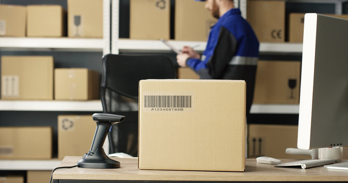 Close up of carton box with bar code is on table at computer in postal office store with parcels. Mail Caucasian courier with invoice and document working on background. Mailman counting boxes.