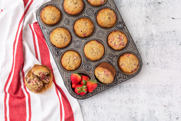 Top View of Homemade Strawberry Muffins in Vintage Muffin Pan; Red and White Kitchen Towel; One Muffin Open on Towel; Fresh Strawberries in One Opening