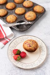 Freshly Baked Homemade Strawberry Muffins ; One Isolated on Plate in Front with Fresh Strawberries; Vintage Muffin Pan in Background; Red and White Kitchen Towel
