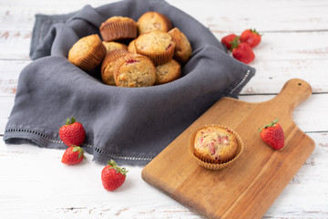 Fototapeta premium Bowl of Freshly Baked Homemade Strawberry Muffins in a Wood Bowl; One Isolated on Wood Cutting Board in Front; Fresh Strawberrries Scattered Around; White Wood Tabletop