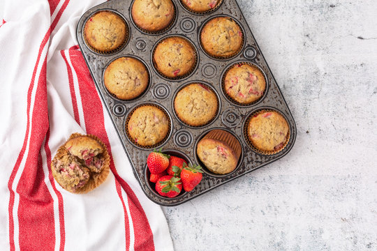 Top View of Homemade Strawberry Muffins in Vintage Muffin Pan; Red and White Kitchen Towel; One Muffin Open on Towel; Fresh Strawberries in One Opening