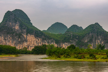 Guilin, China - May 10, 2010: Along Li River. Landscape with green forested karst mountains with brown-black vetical cliffs under dark gray sky. Some trees, a bit of marsh land and Brown water.