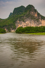 Fototapeta na wymiar Guilin, China - May 10, 2010: Along Li River. Landscape with green forested karst mountains with brown-black vetical cliff under dark gray sky. Some trees, and Brown water up front.