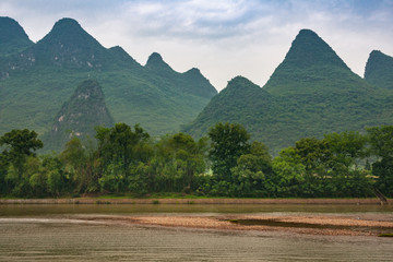 Fototapeta premium Guilin, China - May 10, 2010: Along Li River. Landscape with chain of green forested karst mountain and under blue cloudscape. Green tree belt, Brown water and marsh land up front.