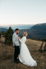 Beautiful couple having a romantic moment on their weeding day, in mountain, sunset.She is in a white wedding dress with a bouquet of sunflowers in hand,groom in a suit. Groom holds bride in his hands