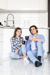 Happy young couple relaxing in kitchen having coffee. Young man and woman sitting on kitchen floor drinking coffee