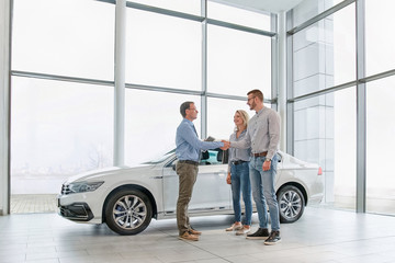 Sales manager helping young couple  choose a new car and shaking hands after a successful car buying in modern car showroom
