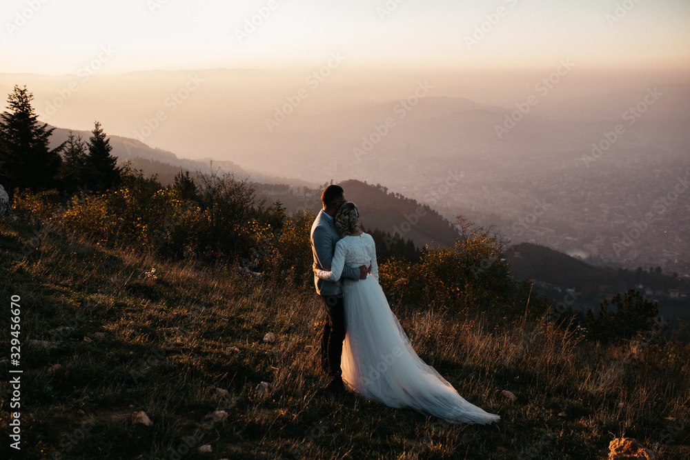Wall mural Beautiful couple having a romantic moment on their weeding day, in mountains at sunset. Bride is in a white wedding dress with a bouquet of sunflowers in hand, groom in a suit. Happy hugging couple. - Wall murals