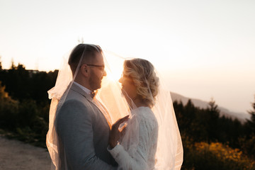 Beautiful couple having a romantic moment on their weeding day, in mountains at sunset. Bride is in a white wedding dress holding bouquet, groom in a suit. Happy hugging couple under the veil. 