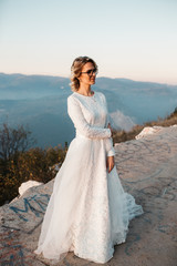 Fototapeta na wymiar Portrait of Beautiful elegant bride in lace wedding dress with long full skirt and long sleeves. Pretty girl in white. Nature, with city in the background, sunset time.