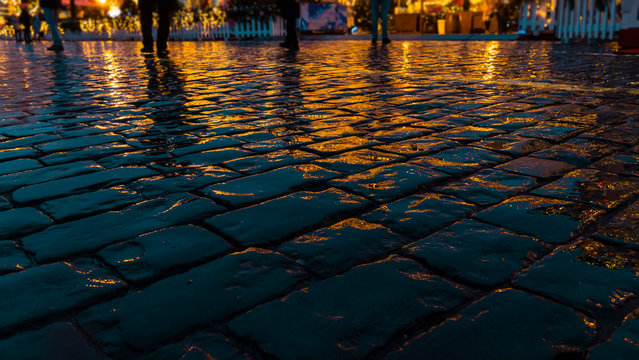 Pavement. Wet paving stones after the rain. Against the background of bright yellow lights of the fair and blurred silhouettes of people . The light reflects off the wet stones.