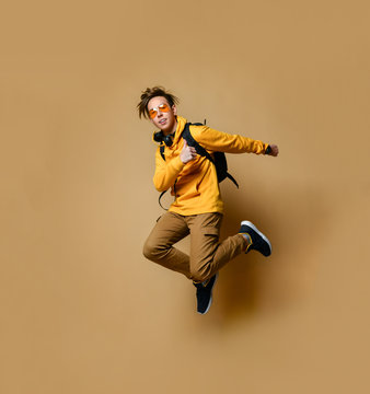 Young teen boy in comfortable clothing, sneakers, sunglasses and backpack jumping and feeling cool over yellow wall background