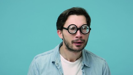 Closeup of comical awkward geek adjusting round diopter eyeglasses, looking around with silly funny grimace, having vision problems, eye health concept. indoor studio shot isolated on blue background