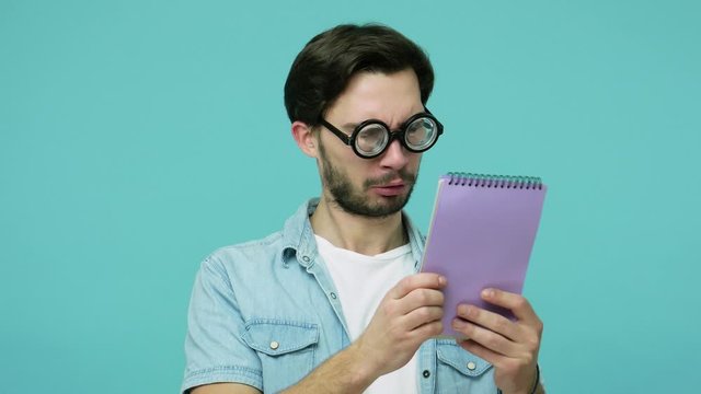Comical weird geek in glasses studying. Clever bearded guy with round diopter eyeglasses reading notebook entries and gazing at camera, checking list-to-do. studio shot isolated on blue background