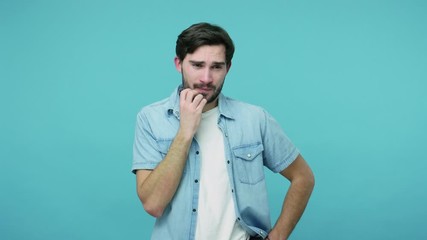 Worried confused guy in jeans shirt rubbing his bristle and thinking over difficult question, expressing doubts, hesitating about difficult choice. indoor studio shot isolated on blue background