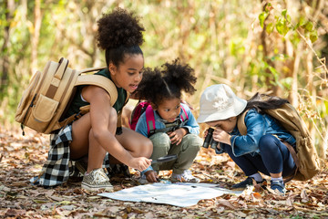 Group of happy pretty little girl hiking together with backpacks and sitting on forest dirt road with looking at the map for exploring the forest. Three kids having fun adventuring in sunny summer day - Powered by Adobe