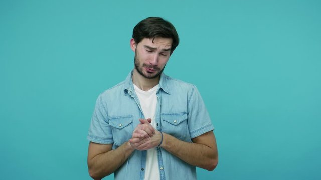 Funny imploring bearded guy in jeans shirt holding hands in prayer gesture and begging, pleading with humble eyes, repeating to say please, urging help. indoor studio shot isolated on blue background