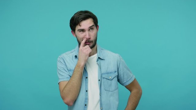 Comical bearded guy in jeans shirt picking dirty nose, pulling out boogers with stupid humorous expression, uncultured bad habit, misconduct concept. indoor studio shot isolated on blue background