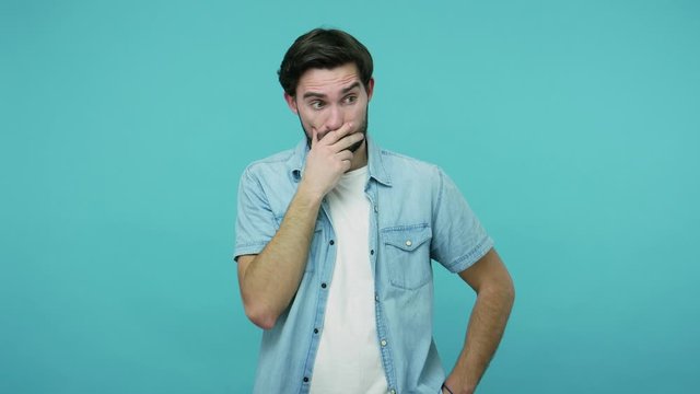 No, I won't say anything. Shocked confused scared bearded guy in jeans shirt covering mouth with hand, looking depressed and amazed by horrible truth secret. studio shot isolated on blue background