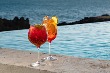 A cold refreshing red and orange bright colored cocktail with ice cubes and orange slices on pool and ocean background