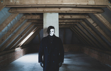 Fototapeta na wymiar A scary guy in white mask with black hair and long dark coat standing in an abandoned attic. Scary and mystical picture. Horror concept.