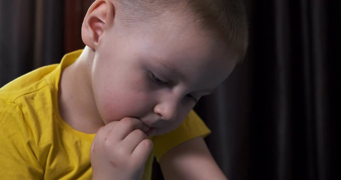 Boy of preschool age lies in bed on his stomach, bites nails on hand, looks at tablet screen, does not take his eyes off, props cheek with hand, close-up, children's room, home furnishings, childhood