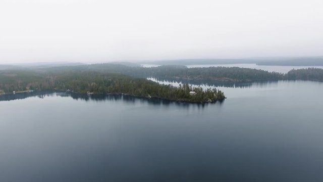 The drone slowly approaches a house in a dense forest on the shore of a large and calm lake on a foggy day (Willard Lake, Ontario, Canada)