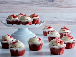 A dozen mini red velvet cupcakes on a white platter and surrounded by more on the table in front of it.  Delicious dessert!