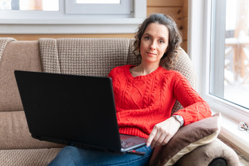 Female works and watches social networks on her computer. An attractive woman with a laptop is sitting on the sofa by the window.
