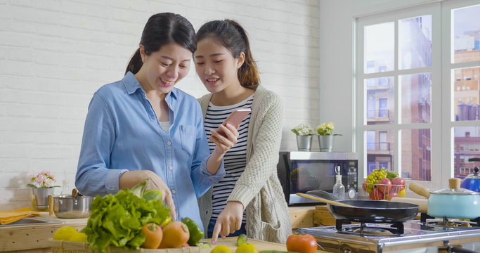 Shot of group of asian japanese female friends cooking in cozy kitchen together. two elegant women roommates discussing process in cooking place. girls holding cellphone checking food material