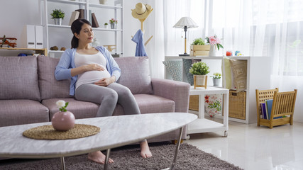 asian japanese pregnant woman sit on grey sofa with her hand on her stomach. Waiting for the birth of unborn baby in belly. young elegant lady in maternity relax in couch in living room at home.