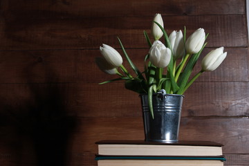 Bunch of white tulips in tin bucket on stack of books against wooden background with copy space. Spring concept. Reading, knowledge, education, decoration