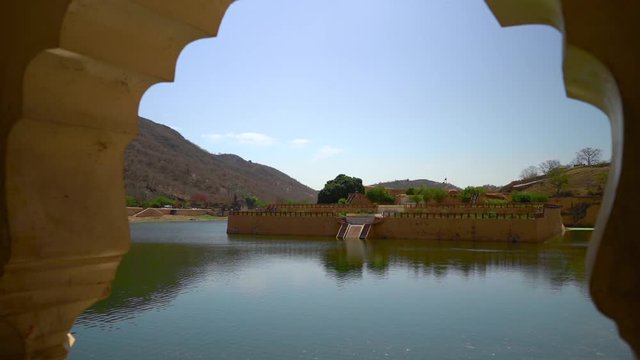 pan in shot through an arched window of the blue green water and kesar kyari bagh island in the maota lake in Amber palace in Jaipur