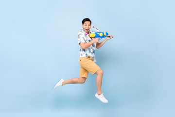 Handsome smiling young Asian man playing with water gun and jumping in studio blue background for Songkran festival in Thailand and southeast Asia