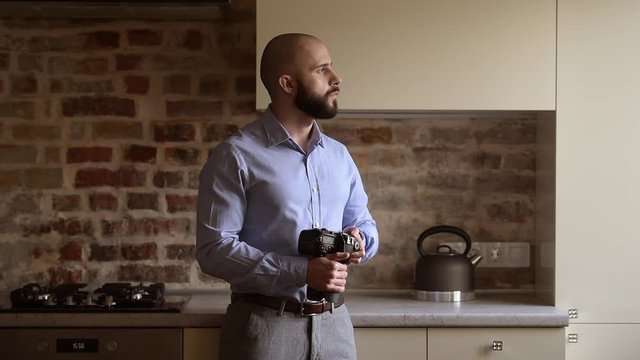 A bald photographer searches for some preferences in his camera then looks to the side. A man with a beard wears two silver earrings, a blue shirt, grey trousers, and a brown leather belt.
