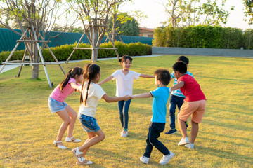 Large group of happy Asian smiling kindergarten kids friends holding hands playing and dancing play roundelay and stand in circle in the park on the green grass on sunny summer day..