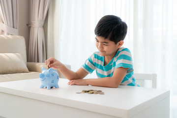 Naklejka premium Little Asian boy insert a coin into blue piggy bank in white table at living room at home for the kid saving, investment money for his business future concept.