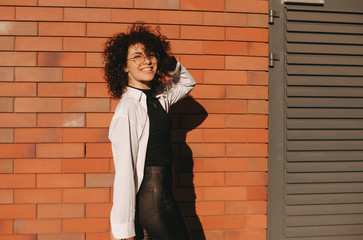 Caucasian businesswoman with nice curly hair and eyeglasses is posing cheerfully on a stone wall