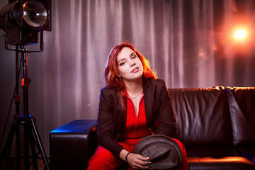 Beautiful young woman with redhair, black hat and red dress on black sofa in the room and nice light behind her