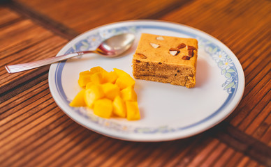 Close up of a piece of gooey chewy vanilla blondie with chopped almond topping served with freshly cut mango pieces on a wooden table mat background. Protein cake recipe and healthy breakfast concept.