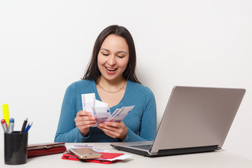 Young cheerful woman holding fan of cash money banknotes, sit and work at desk with pc laptop on white background