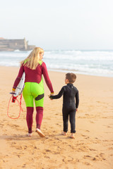 Fototapeta na wymiar Back view of mother and son with surfboard on beach. Rear view of mother and cute little son in wetsuits walking together on sandy beach. Surfing concept