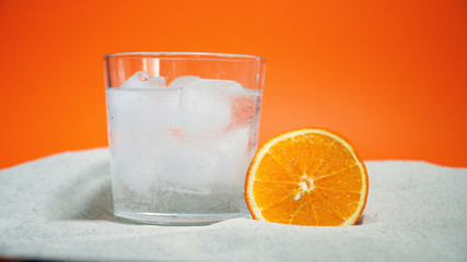 glass with ice and soda water and orange on sand on an orange background