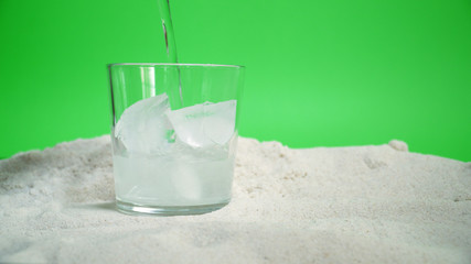 glass with ice and soda water on the sand on a green background