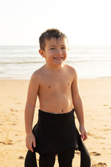Fototapeta premium Adorable child in wetsuit smiling at camera. Portrait of cute happy little boy standing on sandy beach and looking at camera. Childhood and water sport concept