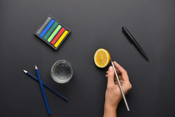 Female hand with stationery, lemon and glass of water on dark background