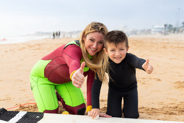 Fototapeta na wymiar Mother and son in wetsuits showing thumbs up. Happy mother and cute little son waxing surfboard and smiling at camera together on sandy sea coast. Surfing concept