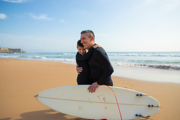 Obraz premium Father and son in wetsuits hugging on beach. Cheerful father and son hugging while spending time together with surfboard on beach. Water sport concept