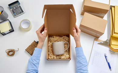 delivery, mail service, people and shipment concept - female hands packing mug to parcel box with straw filler at post office - obrazy, fototapety, plakaty