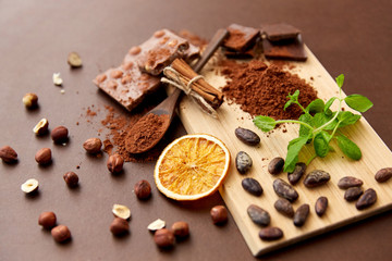 sweets, confectionery and culinary concept - chocolate with hazelnuts, cocoa beans, and powder with dry orange and cinnamon on wooden board with spoon on brown background